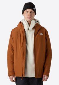 Chaqueta impermeable marrón con capucha ajustable, llevada sobre un polar crema. Presenta cremalleras, puños elásticos y detalles con el logotipo.