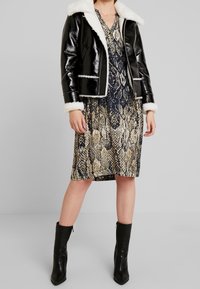 Černá umělá kožená bunda s bílým beránkovým límcem a obšíváním, nošená přes šaty s hadím vzorem v béžové a námořnické modři. Černé kotníkové boty dotvářejí vzhled.