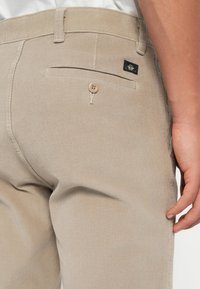 Pantalon en velours côtelé beige au design ajusté, avec une poche arrière à fermeture par bouton et une petite étiquette en tissu sur la ceinture.