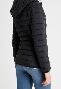 Černá zateplená bunda s kapucí, s horizontálním prošíváním, měkkou texturou a přiléhavým střihem, kombinovaná s modrými džíny.