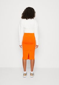 Maglione bianco corto con maniche lunghe abbinato a una gonna midi aderente arancione brillante con uno spacco posteriore, indossato con scarpe da ginnastica multicolori.