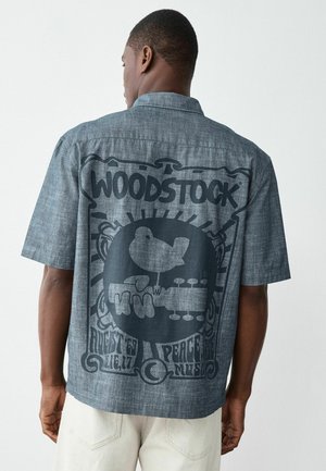 Muž nosi modré tričko s krátkym rukávom s veľkým čiernym grafikým motívom Woodstock zobrazujúcim vtáka na gitare a textom o hudobnom festivale.