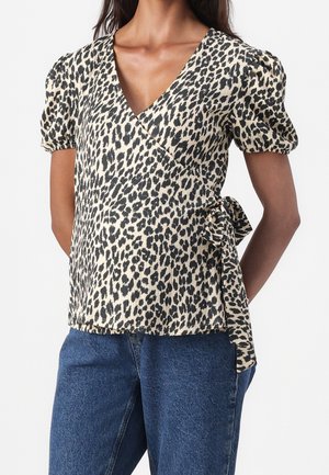 Kvinde iført en beige og sort leopardmønstret slå-om-bluse med puffede korte ærmer og bånd i siden, kombineret med blå denimjeans.