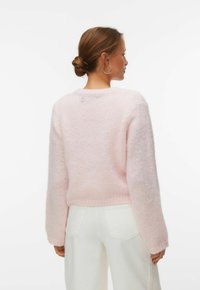 Femme aux cheveux bruns attachés en chignon, portant un pull rose clair duveteux et un pantalon blanc, vue de dos sur fond blanc uni.
