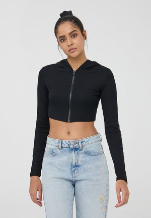 Hoodie nera corta con maniche lunghe, chiusura frontale con zip, texture a coste. Abbinato a jeans chiari strappati, con vita alta.