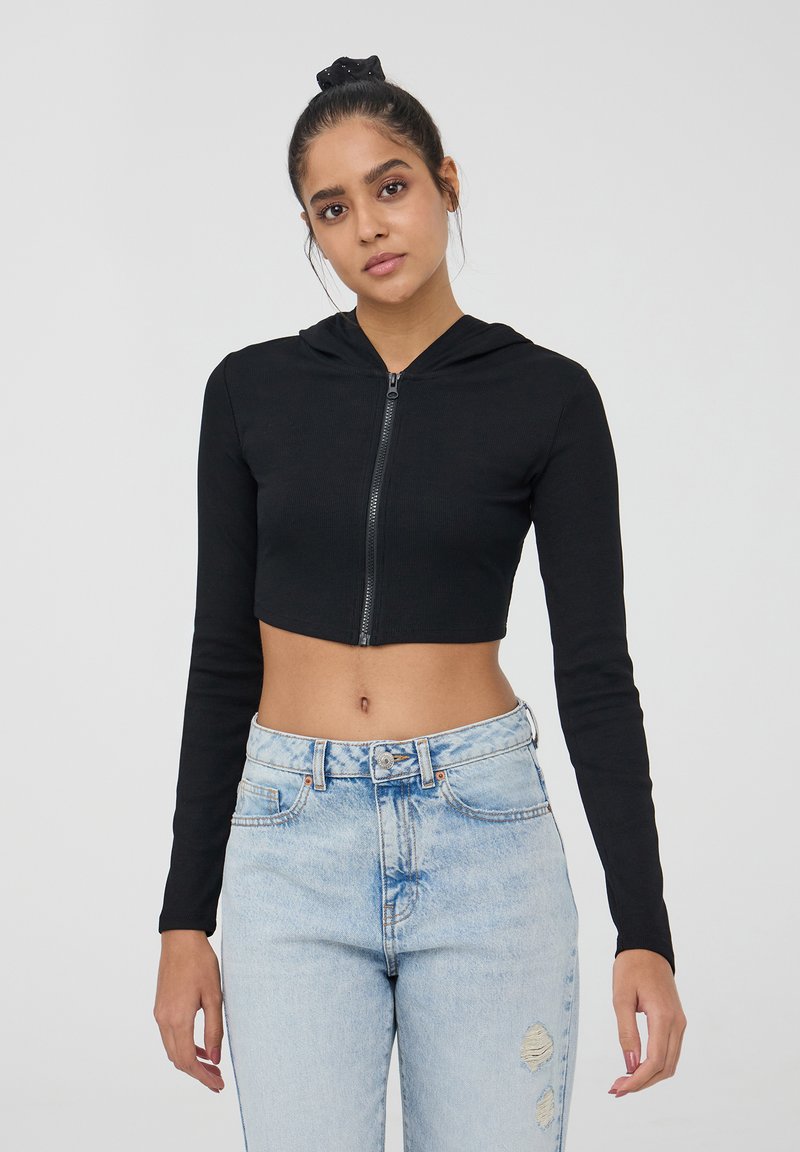 Hoodie nera corta con maniche lunghe, chiusura frontale con zip, texture a coste. Abbinato a jeans chiari strappati, con vita alta.
