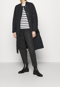 Cappotto lungo trapuntato nero con colletto rotondo, abbinato a una camicia a righe bianche e navy, jeans scuri e stivaletti neri.