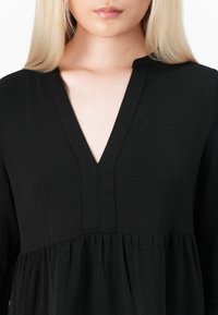 Svart, texturerad tunika med V-ringning och plisserad design. Har subtila sömmar och en lös, svävande silhuett. Materialet verkar vara lätt.