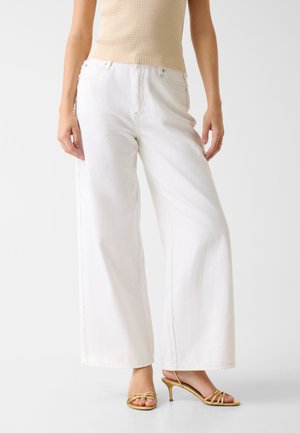 Femme portant un jean blanc taille haute à jambes larges, un top beige sans manches côtelé, et des sandales à talons dorées à brides, debout devant un fond uni.