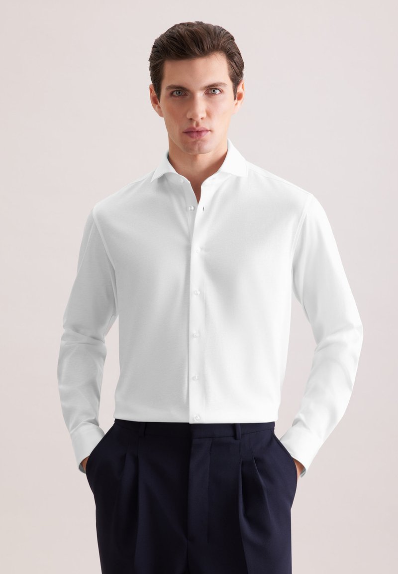 Jeune homme portant une chemise blanche cintrée à boutons, rentrée dans un pantalon plissé foncé, debout les mains dans les poches devant un fond uni.