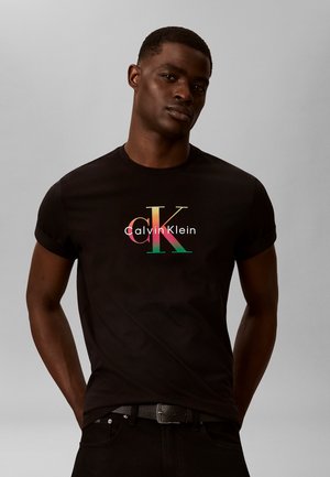 Άντρας που φοράει μαύρο κοντομάνικο Calvin Klein με πολύχρωμο λογότυπο «cK», στέκεται με τα χέρια πίσω από την πλάτη σε απλό γκρι φόντο.