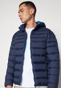 Tamnoplava puffa jakna s kapuljačom, sa kvadratnim uzorkom i plavim naglascima, prednje zatvaranje na zip, te lagani, izolirani materijal.