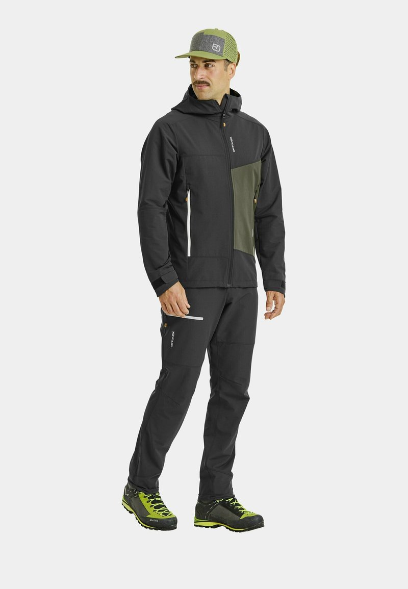 Black jacket with olive green accents, zip front, elastic cuffs; matching black pants; green and grey cap; rugged outdoor shoes with yellow details.
