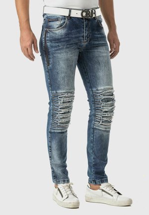 Blaue Denim-Jeans mit distressed Details, aufgerissenen Akzenten an den Knien, schlankem Schnitt und einem weißen Gürtel über einem enganliegenden weißen T-Shirt.