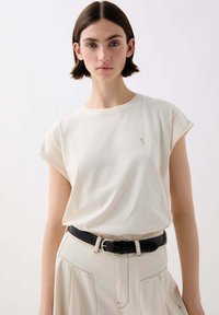 T-shirt crema a maniche corte con maniche arrotolate e un piccolo logo ricamato, abbinato a una gonna beige a vita alta e texturizzata con cintura nera.