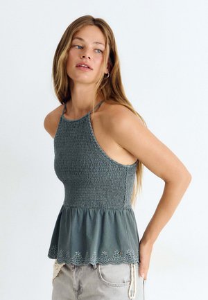 Femme aux longs cheveux portant un haut sans manches smocké bleu sarcelle avec une broderie à l'ourlet et un short en jean clair.