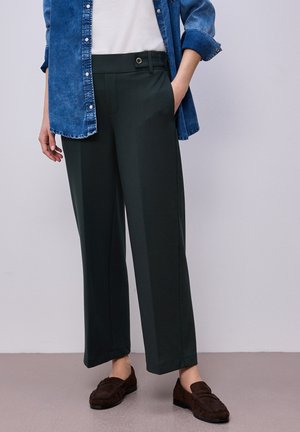 Person iført mørkegrønne vide bukser, hvid top, blå denim skjorte med knapper og brune loafers, stående med den ene hånd i lommen.