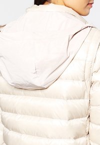 Nafoukaná béžová bunda s hladkou texturou, vybavená kapucí a horizontálními prošívanými sekcemi. Materiál vypadá lehký a voděodolný.