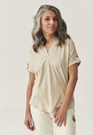 Beige, lyhythihainen polo-paita, jossa on V-kaulus ja käännetyt hihansuut. Pehmeä kangas, rento malli, yhdistetty valkoisiin housuihin, joissa on brodeerattu yksityiskohta.
