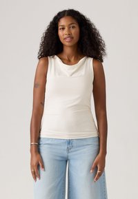 Frau mit lockigem Haar, die ein ärmelloses weißes Oberteil und helle blaue Jeans trägt und vor einem schlichten hellen Hintergrund steht.