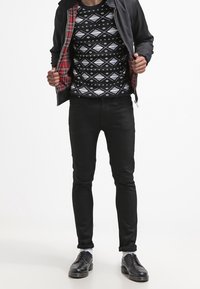 Veste noire avec doublure en tartan rouge, pull noir à motifs, jean noir slim et chaussures noires avec chaussettes blanches.