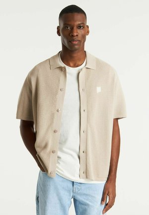 Beige gestricktes kurzärmeliges Hemd mit Kragen, ausgestattet mit Knopfverschluss und einer strukturierten Musterung. Getragen über einem schlichten weißen T-Shirt.