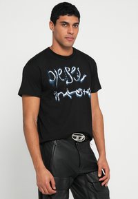 Μαύρο βαμβακερό T-shirt με μπλε και λευκή γραφική σχεδίαση κειμένου. Κοντά μανίκια, στρογγυλή λαιμόκοψη, φορεμένο με μαύρο δερμάτινο παντελόνι.