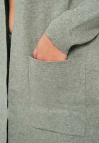 Ανοιχτό γκρι καπέλο από βελούδο με υφή, διαθέτει μακριά μανίκια και μια μεγάλη τετράγωνη μπροστινή τσέπη στη δεξιά πλευρά.