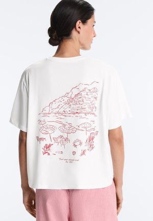 Donna con capelli scuri raccolti in uno chignon che indossa una maglietta bianca con un disegno a linee rosse di una scena di spiaggia, ombrelloni, sedie, granchio e paese sulla collina sulla schiena.