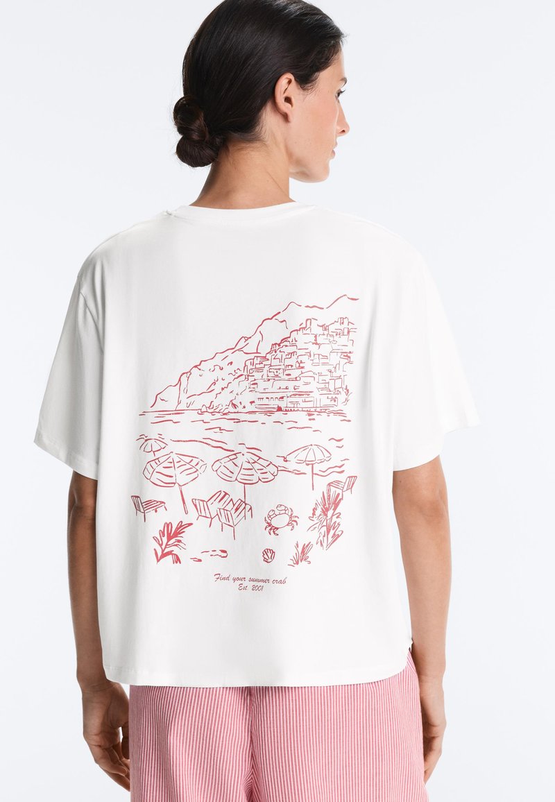 Femme aux cheveux foncés en chignon portant un t-shirt blanc avec un dessin rouge au trait représentant une scène de plage, des parasols, des chaises, un crabe et un village sur une colline au dos.