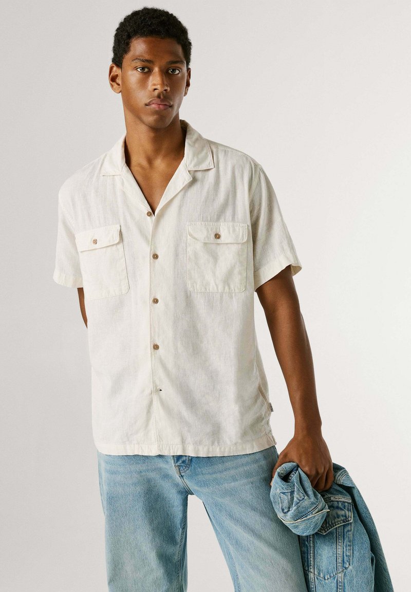 Jeune homme debout portant une chemise légère à manches courtes boutonnée et un jean, tenant une veste en jean pliée dans sa main droite.