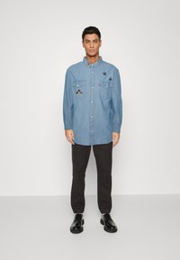 Light blue denim shirt with two chest pockets, embroidered mushrooms and insects, paired with black striped pants and black shoes.