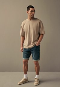 Muškarac u bež oversized majici, s odrezanim trapericama, bijelim čarapama i bež tenisicama, stoji uz jednostavan bež zid.