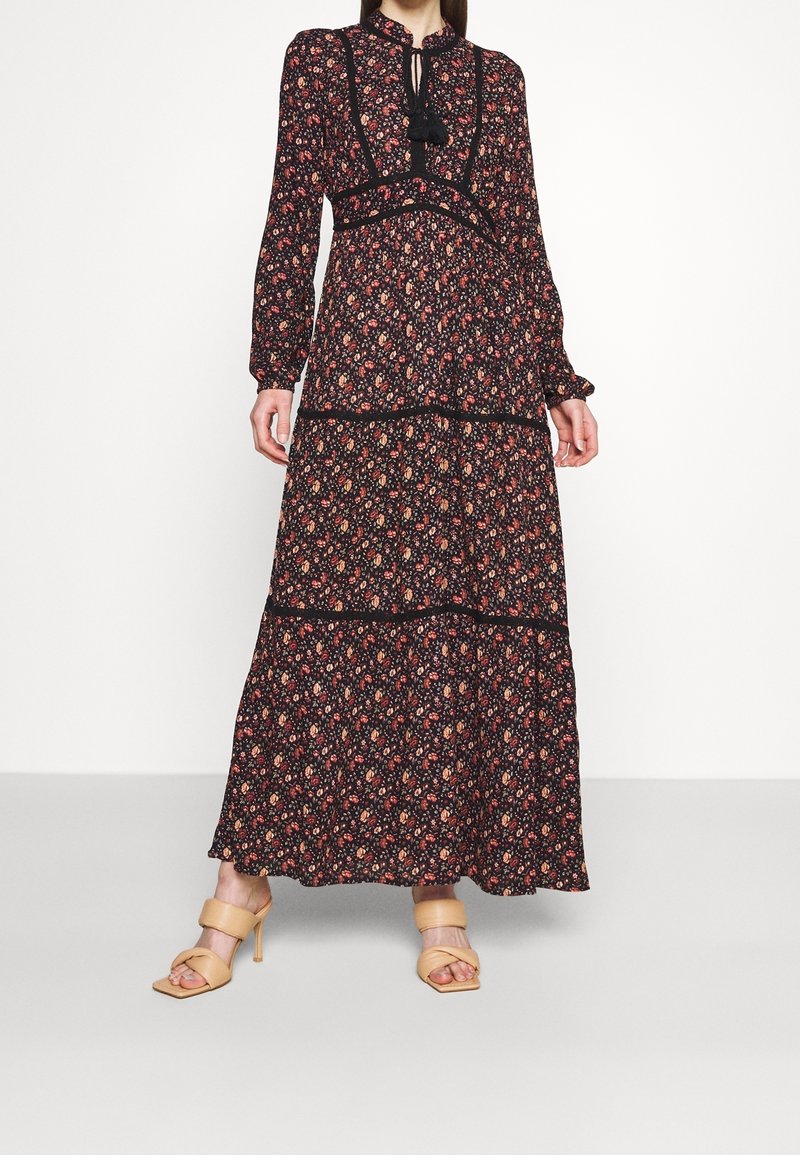 Femme portant une longue robe noire à fleurs avec des manches longues, un bord noir, et des sandales à talons beiges, debout devant un fond blanc uni.