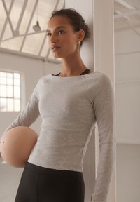 Langärmliges, hellgraues Oberteil mit gerippter Textur und rundem Ausschnitt, kombiniert mit schwarzen, figurbetonten Leggings. Model hält einen hellfarbenen, runden Gegenstand.