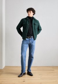 Veste verte texturée avec poches avant, col roulé noir, jeans bleu clair et chaussures marron ; debout devant un fond gris clair.