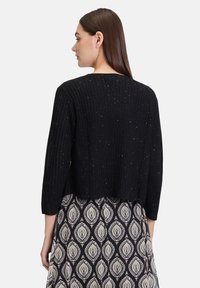 Cardigan noir court avec texture côtelée et éclats argentés. Présente des manches trois-quarts et un col rond.