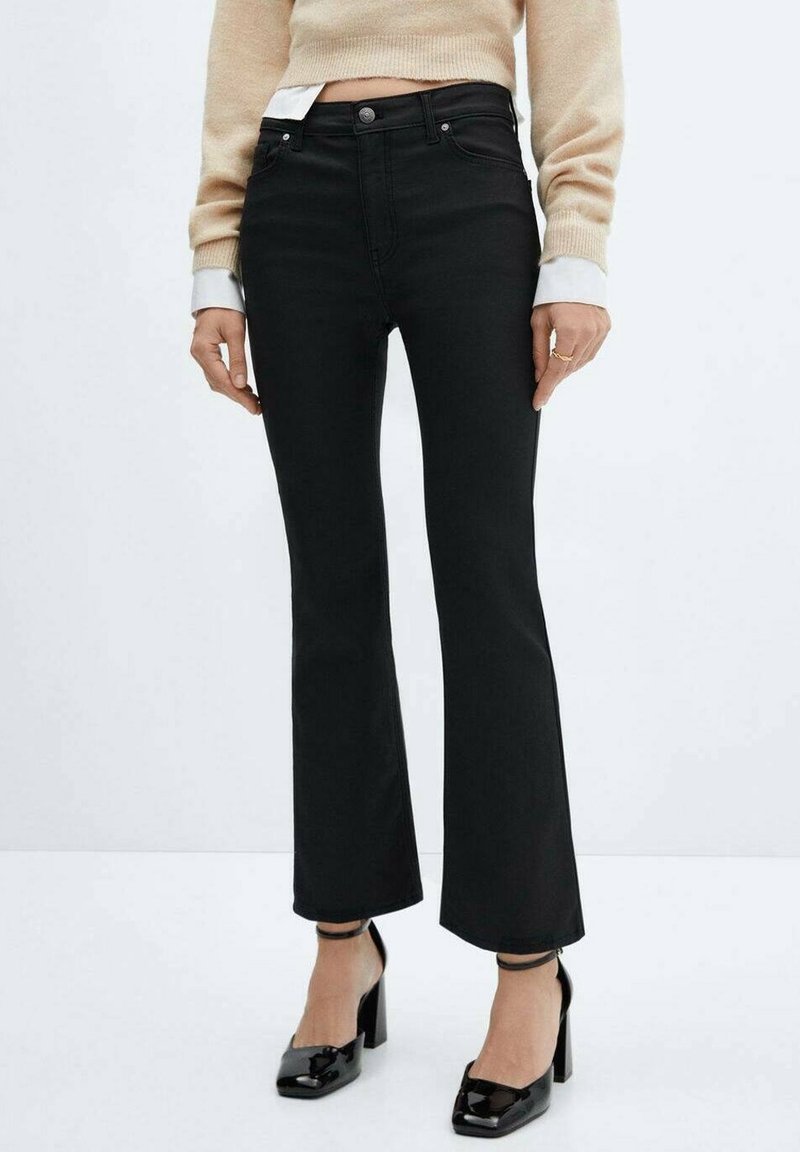 Femme portant un jean noir évasé taille haute, un pull court beige et des escarpins vernis noirs à brides à la cheville.
