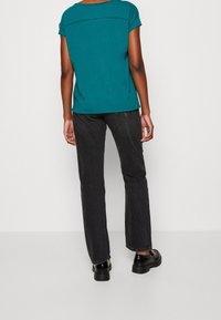 Top corto teal a maniche corte con scollatura rotonda, abbinato a pantaloni jeans dritti scuri e scarpe nere. Tessuto liscio e design informale.