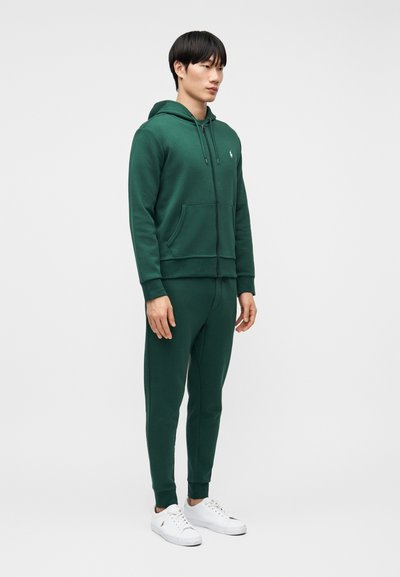 Άνδρας που στέκεται φορώντας σκούρο πράσινο φούτερ με φερμουάρ, ταιριαστό παντελόνι φόρμας και λευκά αθλητικά παπούτσια μπροστά σε απλό ανοιχτό γκρι φόντο.