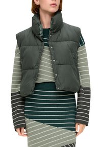 Gepolsterte grüne Weste mit hohem Kragen, Druckknopfverschluss und seitlicher Reißverschlusstasche, kombiniert mit einem mehrfarbigen grauen und grünen Strickkleid.