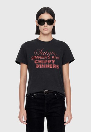 Person wearing black sunglasses and a black t-shirt with red text reading "Saints, Sinners and Chippy Dinners" standing against a plain background.