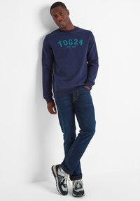 Navy blue sweatshirt with green "TOG24" text, paired with denim jeans and black athletic shoes with grey and green accents.