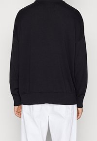 Schwarzer, übergroßer Pullover mit rundem Halsausschnitt, geripptem Saum und abgesenkten Schultern, vor einem einfarbigen Hintergrund präsentiert.