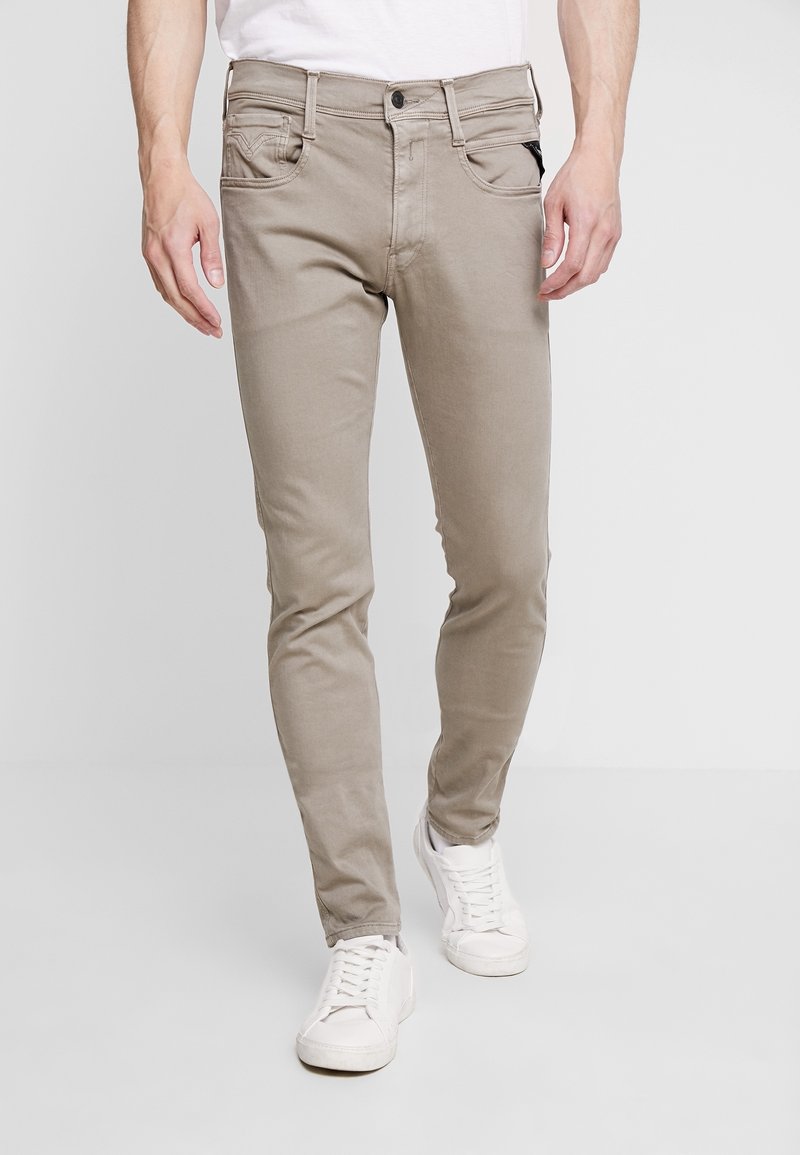 Vaqueros de denim ajustados color beige con cinco bolsillos, cierre de botón y ligero estiramiento para mayor comodidad; combinados con zapatillas deportivas blancas.