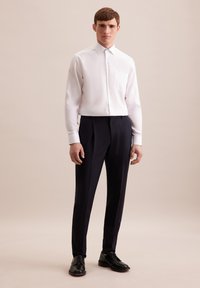 Camicia bianca a maniche lunghe con taschino, abbinata a pantaloni su misura blu scuro. Il modello indossa scarpe nere, in piedi su uno sfondo neutro.