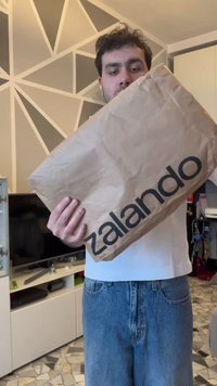 Άντρας κρατάει ένα μεγάλο καφέ πακέτο Zalando σε ένα δωμάτιο με γεωμετρική τέχνη στους τοίχους και πλακάκια στο πάτωμα.