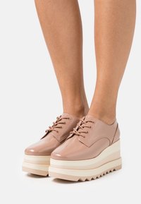 Chaussures compensées en cuir verni nude brillant, avec bout rond, lacets sur le devant, et plateforme superposée à semelle dentelée pour une meilleure traction.