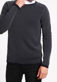 Pull à manches longues bleu marine avec motif à pois blancs, col rond et poignets côtelés. Le tissu semble tricoté, offrant une finition texturée.