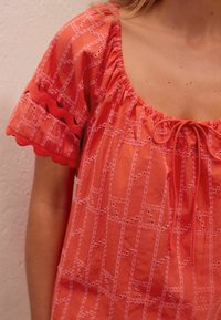 Rode top met een brede halslijn, complexe witte borduursels, scalloped mouwen en een strikdetail aan de voorkant. Lichtgewicht stof met een gestructureerd patroon.